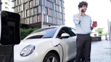 Takım elbiseli, hırslı bir işadamı elektrik kablosu ve yenilenebilir enerji enerjili bir elektrikli araçla şarj istasyonunda dururken elektrikli aracına yaslanıyor..