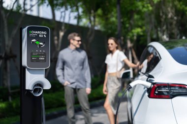 Genç çift yazın yeşil sürdürülebilir şehir bahçesinde EV elektrikli araba ile seyahat ederken, yeşil temiz yeniden şarj edilebilir elektrikli araç iç organları ile kentsel sürdürülebilirlik yaşam tarzını gösteriyor.