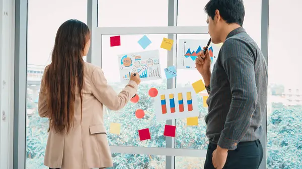 Business people work on project planning board in office and having conversation with coworker friend to analyze project development . They use sticky notes posted on glass to make it organized. Jivy