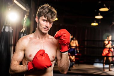 Boksör gömleksiz, beyaz erkek boksör savunma pozisyonu almış kırmızı eldiven giymiş arka planda ring ve boks ekipmanıyla dövüşmeye ve yumruk atmaya hazır. Impetus