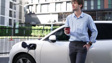 İlerici işadamı, şehir merkezinde dolaşmadan önce şarj cihazından elektrikli arabasına şarj prizi takıyor. Çevre dostu şarj edilebilir araba, sürdürülebilir ve temiz enerjiyle çalışıyor..
