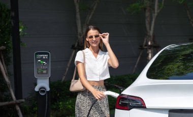 Genç bir kadın yazın yeşil sürdürülebilir şehir bahçesinde EV elektrikli arabasıyla seyahat ediyor. Kentsel sürdürülebilirlik yaşam tarzı yeşil temiz yeniden şarj edilebilir enerji elektrikli BEV araç iç organları