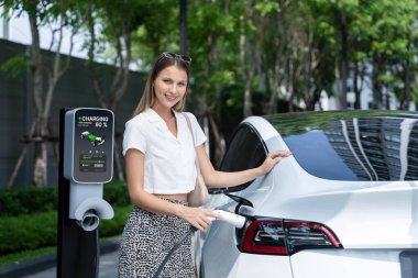 Genç bir kadın yazın yeşil sürdürülebilir şehir bahçesinde EV elektrikli arabasıyla seyahat ediyor. Kentsel sürdürülebilirlik yaşam tarzı yeşil temiz yeniden şarj edilebilir enerji elektrikli BEV araç iç organları