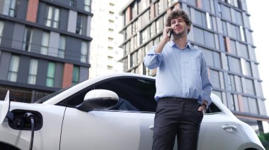 Takım elbiseli, hırslı bir işadamı elektrik kablosu ve yenilenebilir enerji enerjili bir elektrikli araçla şarj istasyonunda dururken elektrikli aracına yaslanıyor..
