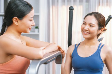 Enerjik ve güçlü atletik Asyalı kadın evde spor arkadaşı ya da antrenörü ile birlikte eliptik koşu makinesiyle koşuyor. Formda fiziğin takibi ve sağlıklı yaşam tarzına bağlılık. Kuvvetli