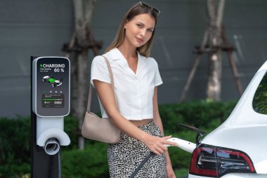 Genç bir kadın yazın yeşil sürdürülebilir şehir bahçesinde EV elektrikli arabasıyla seyahat ediyor. Kentsel sürdürülebilirlik yaşam tarzı yeşil temiz yeniden şarj edilebilir enerji elektrikli BEV araç iç organları