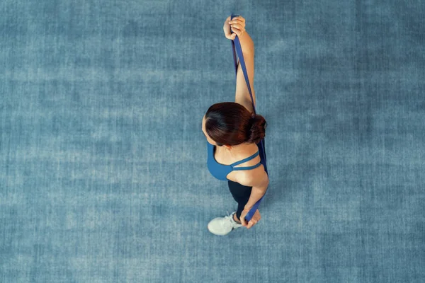 Üst görüşlü enerjik kadın halı yüzeyi arka planında egzersiz yapıyor, direnç sporu bandını esnetiyor. Genç atletik Asyalı kadın gücü ve dayanıklılığı evde antrenman rutini olarak kullanılıyor..