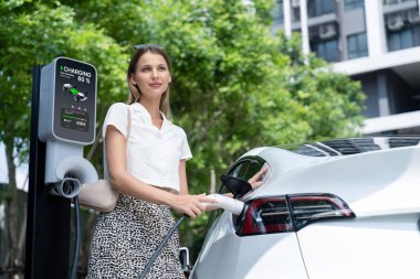 Genç bir kadın yazın yeşil sürdürülebilir şehir bahçesinde EV elektrikli arabasıyla seyahat ediyor. Kentsel sürdürülebilirlik yaşam tarzı yeşil temiz yeniden şarj edilebilir enerji elektrikli BEV araç iç organları