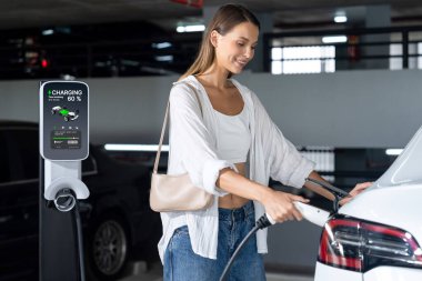 Genç bir kadın EV elektrikli arabasıyla alışveriş merkezinin otoparkına gidiyor şehir merkezinde elektrik iç organlarının yeşil temiz şarj edilebilir enerji ile sürdürülebilirlik yaşam tarzını gösteriyor.