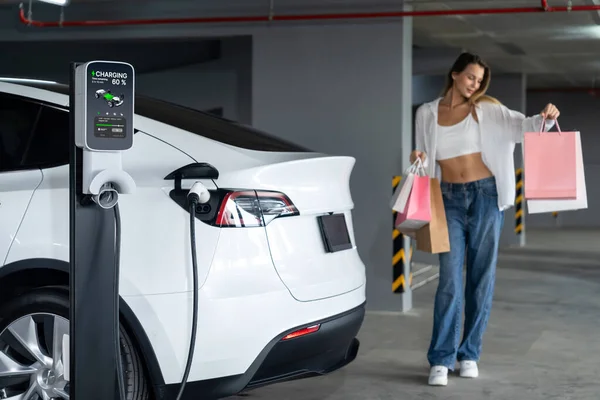 若い女性はEv電気自動車で街の中心部の駐車場に充電し 電気自動車の旅館の緑のきれいな再充電可能エネルギーによって都市の持続可能性のライフスタイルを示しています — ストック写真