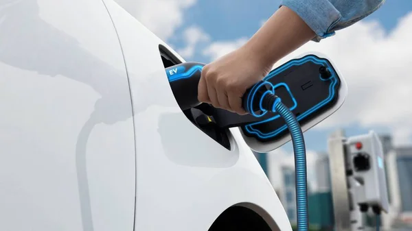 Asian man insert EV charger and recharge his electric car from future charging station with blurred background of cityscape. Smart and futuristic sustainable energy infrastructure. Peruse