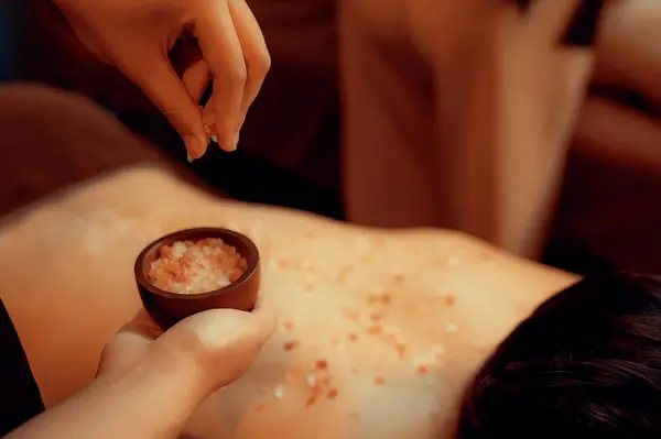 Closeup man customer having exfoliation treatment in luxury spa salon with warmth candle light ambient. Salt scrub beauty treatment in Health spa body scrub. Quiescent