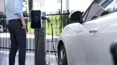 İlerici işadamı, şehir merkezinde dolaşmadan önce şarj cihazından elektrikli arabasına şarj prizi takıyor. Çevre dostu şarj edilebilir araba, sürdürülebilir ve temiz enerjiyle çalışıyor..