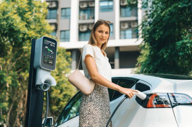 Genç bir kadın yazın yeşil sürdürülebilir şehir bahçesinde EV elektrikli arabasıyla seyahat ediyor. Kentsel sürdürülebilirlik yaşam tarzı yeşil temiz yeniden şarj edilebilir enerji elektrikli BEV araç iç organları
