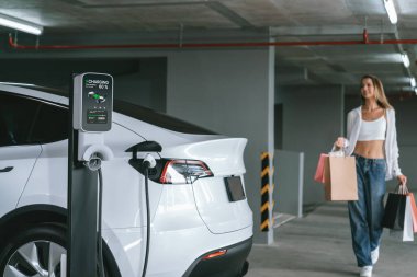 Genç bir kadın EV elektrikli arabasıyla alışveriş merkezinin otoparkına gidiyor şehir merkezinde elektrik iç organlarının yeşil temiz şarj edilebilir enerji ile sürdürülebilirlik yaşam tarzını gösteriyor.