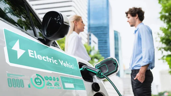 充電ステーションから電気自動車を充電した後に待っているビジネスマンは 駐車場でスマートバッテリーステータスホログラムを表示します 環境に優しいEv車は 都市生活の中で代替エネルギーを使用します Peruse — ストック写真