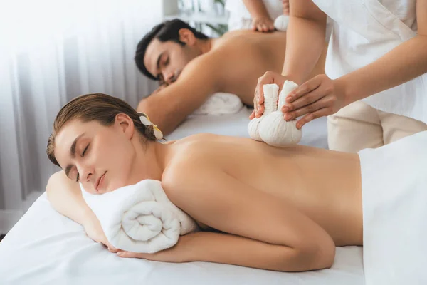 Hot herbal ball spa massage body treatment, masseur gently compresses herb bag on couple customer body. Tranquil and serenity of aromatherapy recreation in day lighting ambient at spa salon. Quiescent