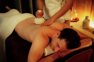 Sıcak bitkisel top spa masaj vücut tedavisi, masör erkek vücuduna nazikçe şifalı bitki torbası bastırıyor. Aromaterapi tesisinin sakinliği ve dinginliği kaplıca salonundaki mumların sıcacık ışığında. Sessiz.