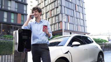 Takım elbiseli, hırslı bir işadamı elektrik kablosu ve yenilenebilir enerji enerjili bir elektrikli araçla şarj istasyonunda dururken elektrikli aracına yaslanıyor..