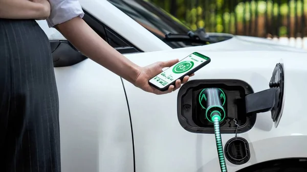 スマートフォンのディスプレイバッテリーステータスインターフェースをスマートEvモバイルアプリケーションで保持するビジネスウーマンと 駐車場の充電ステーションから電気を充電するEvカー Peruse — ストック写真