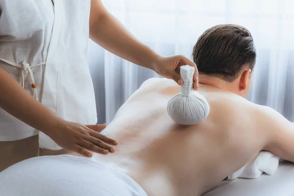 Hot herbal ball spa massage body treatment, masseur gently compresses herb bag on man body. Tranquil and serenity of aromatherapy recreation in day lighting ambient at spa salon. Quiescent