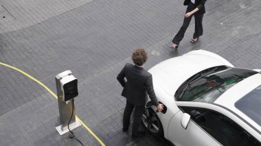İlerici iş adamı ve iş kadını şehir merkezinde dolaşmadan önce şarj istasyonuna bağlı elektrikli arabaya yaslanıyor. Çevre dostu şarj edilebilir araba temiz enerjiyle çalışıyor..