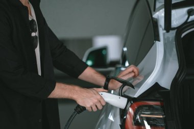 Genç adam EV elektrikli arabasıyla alışveriş merkezinin otoparkına gidiyor şehir merkezindeki şarj oluyor yeşil, temiz, yeniden şarj edilebilir elektrikli araç iç organları ile kentsel sürdürülebilirlik yaşam tarzını gösteriyor