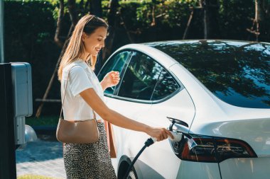 Genç bir kadın yazın yeşil sürdürülebilir şehir bahçesinde EV elektrikli arabasıyla seyahat ediyor. Kentsel sürdürülebilirlik yaşam tarzı yeşil temiz yeniden şarj edilebilir enerji elektrikli BEV araç iç organları