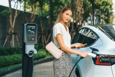 Genç bir kadın yazın yeşil sürdürülebilir şehir bahçesinde EV elektrikli arabasıyla seyahat ediyor. Kentsel sürdürülebilirlik yaşam tarzı yeşil temiz yeniden şarj edilebilir enerji elektrikli BEV araç iç organları