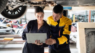 Kaldırılmış arabanın altında birlikte çalışan iki araç tamircisi, dizüstü bilgisayarla araba teftişi yapıyor. Üniformalı otomotiv teknisyeni dikkatle tanısal sorun giderme yapıyor. Panorama Oxus