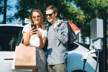 Genç çift yazın yeşil sürdürülebilir şehir bahçesinde EV elektrikli araba ile seyahat ederken, yeşil temiz yeniden şarj edilebilir elektrikli araç iç organları ile kentsel sürdürülebilirlik yaşam tarzını gösteriyor.