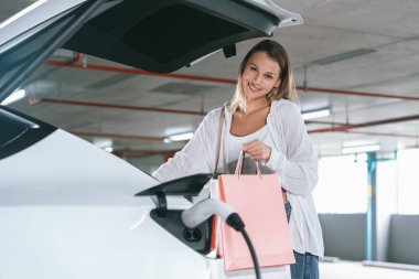 Genç bir kadın EV elektrikli arabasıyla alışveriş merkezinin otoparkına gidiyor şehir merkezinde elektrik iç organlarının yeşil temiz şarj edilebilir enerji ile sürdürülebilirlik yaşam tarzını gösteriyor.