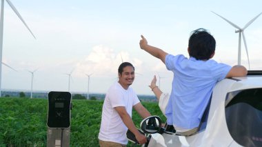 İlerici mutlu aile kavramı elektrikli araçla rüzgar çiftliğinde eğleniyor. Rüzgar türbini jeneratöründen gelen temiz yenilenebilir enerji ile şarj edilen elektrikli araç..