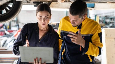 Kaldırılmış arabanın altında birlikte çalışan iki araç tamircisi, dizüstü bilgisayarla araba teftişi yapıyor. Üniformalı otomotiv teknisyeni dikkatle tanısal sorun giderme yapıyor. Panorama Oxus