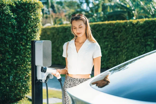 若い女性は夏に緑の持続可能な都市の屋外庭で充電するEv電気自動車で旅行します 電動Bev車両旅館のグリーンクリーン再充電可能エネルギーによる都市持続可能性ライフスタイル — ストック写真