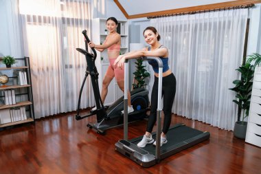 Enerjik ve güçlü atletik Asyalı kadın evde spor arkadaşı ya da antrenörü ile birlikte eliptik koşu makinesiyle koşuyor. Formda fiziğin takibi ve sağlıklı yaşam tarzına bağlılık. Kuvvetli