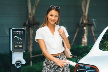 Genç bir kadın yazın yeşil sürdürülebilir şehir bahçesinde EV elektrikli arabasıyla seyahat ediyor. Kentsel sürdürülebilirlik yaşam tarzı yeşil temiz yeniden şarj edilebilir enerji elektrikli BEV araç iç organları