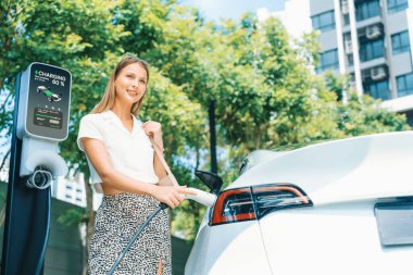 Genç bir kadın yazın yeşil sürdürülebilir şehir bahçesinde EV elektrikli arabasıyla seyahat ediyor. Kentsel sürdürülebilirlik yaşam tarzı yeşil temiz yeniden şarj edilebilir enerji elektrikli BEV araç iç organları