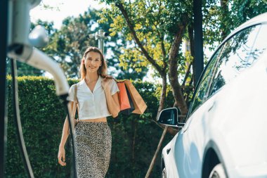 Genç bir kadın yazın yeşil sürdürülebilir şehir bahçesinde EV elektrikli arabasıyla seyahat ediyor. Kentsel sürdürülebilirlik yaşam tarzı yeşil temiz yeniden şarj edilebilir enerji elektrikli BEV araç iç organları