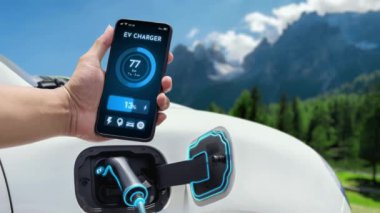 Akıllı telefonlu bir iş adamı akıllı EV mobil uygulama ile batarya durumu arayüzünü tutarken EV otomobili de doğal dağ arka planına sahip şarj istasyonundan elektrik yüklüyor. İncele