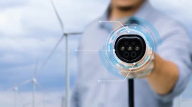 Kameranın önüne odaklanmış EV şarj cihazı fütüristik akıllı dijital EV batarya durum hologramını gösteriyor. EV araba şarjı, ekolojik sürdürülebilir güç için rüzgar türbininin yenilenebilir enerjisini kullanıyor. İnceleyin