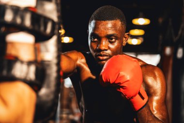 Afrika kökenli siyah boksör antrenöre yumruk atıyor ya da spor salonunda boks çantası olarak boks eldiveni takıyor. Profesyonel boks maçı için güç ve dayanıklılık eğitimi. Impetus