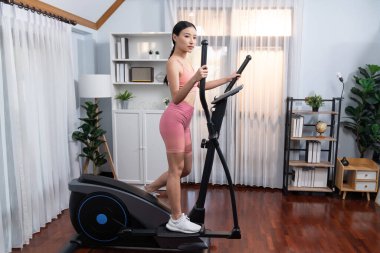 Enerjik ve güçlü atletik Asyalı kadın evde eliptik koşu makinesiyle koşuyor. Formda fiziğin takibi ve ev çalışması ve eğitimle sağlıklı yaşam tarzına bağlılık. Kuvvetli