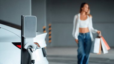 Genç bir kadın EV elektrikli arabasıyla alışveriş merkezinin otoparkına gidiyor şehir merkezinde elektrik iç organlarının yeşil temiz şarj edilebilir enerji ile sürdürülebilirlik yaşam tarzını gösteriyor.