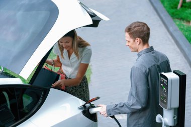 Genç çift yazın yeşil sürdürülebilir şehir bahçesinde EV elektrikli araba ile seyahat ederken, yeşil temiz yeniden şarj edilebilir elektrikli araç iç organları ile kentsel sürdürülebilirlik yaşam tarzını gösteriyor.