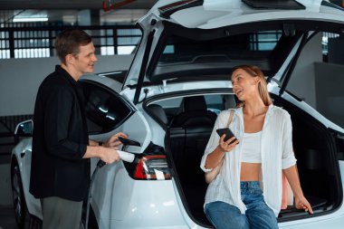 Genç çift EV elektrikli arabasıyla alışveriş merkezinin otoparkına gidiyor şehir merkezindeki şarj oluyor yeşil temiz şarj edilebilir elektrikli araç iç organları ile kentsel sürdürülebilirlik yaşam tarzını gösteriyor