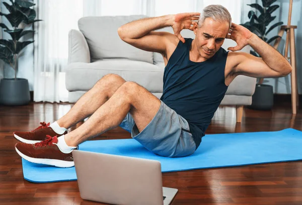Athletic and sporty senior man doing crunch on the exercising mat while watching online exercising session at home exercise as concept of healthy fit body lifestyle after retirement. Clout