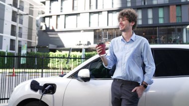 İlerici işadamı, şehir merkezinde dolaşmadan önce şarj cihazından elektrikli arabasına şarj prizi takıyor. Çevre dostu şarj edilebilir araba, sürdürülebilir ve temiz enerjiyle çalışıyor..