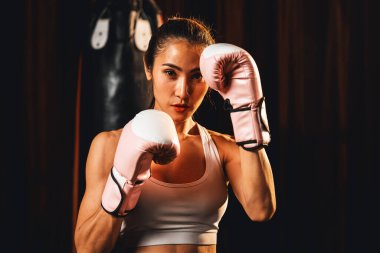 Asya 'lı kadın Muay Thai boksörü kamera önünde boksör yumruğu. Arka planda boks ekipmanıyla spor salonunda poz vermeye hazır. Kararlı gözler ve meydan okumaya hazır olun. Impetus