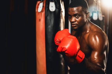 Boksör üstsüz, Afro-Amerikan siyah boksör savunma pozisyonu almış kırmızı eldiven giymiş, arka planda boks ekipmanları ve kum torbalarıyla spor salonunda dövüşmeye ve yumruk atmaya hazır. Impetus